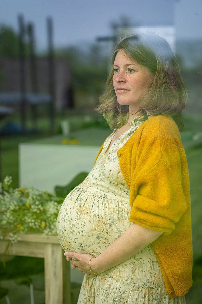 Zwangere vrouw met handen op de buik kijkt dromerig door het raam.