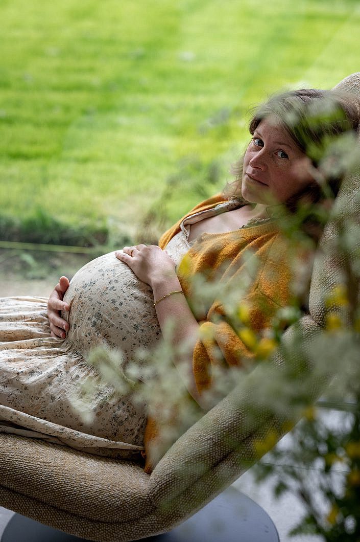 Zwangere vrouw in zetel kijkt recht in de lens door bloemen.