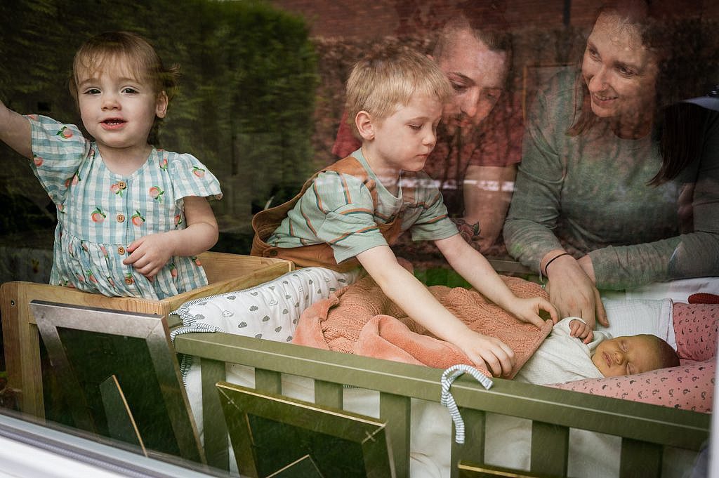 Gezin door het raam met baby uit Lievegem in babybedje.