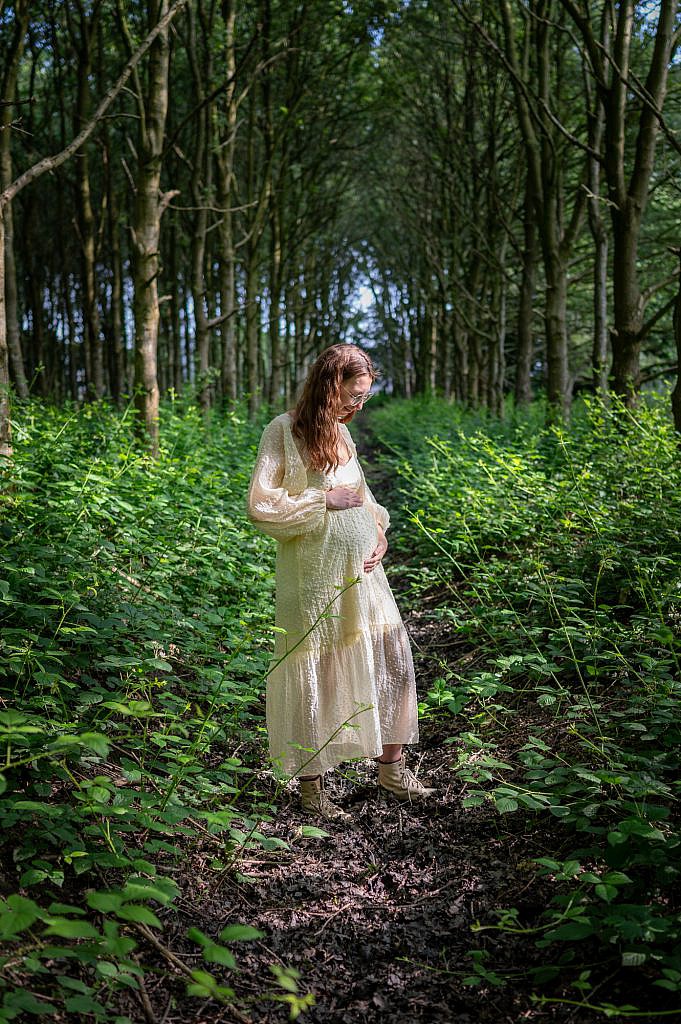 Zwangerschapsfotoshoot van zwangere vrouw in wit kleedje houdt haar buik vast en kijkt ernaar.