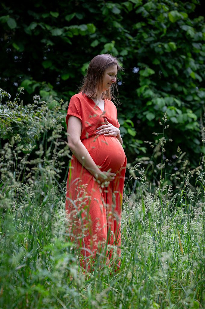 Zwangerschapsfotoshoot van zwangere vrouw in rood kleedje tussen het gras.