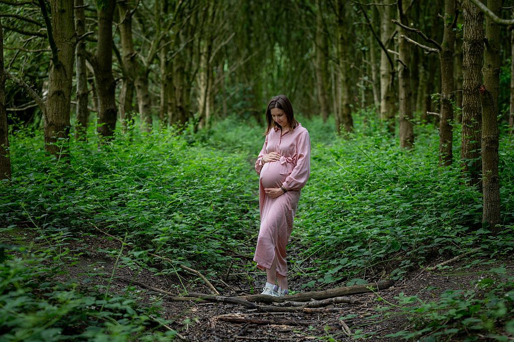 Zwangerschapsfotoshoot van vrouw met roze kleedje houdt haar buik vast in het bos.