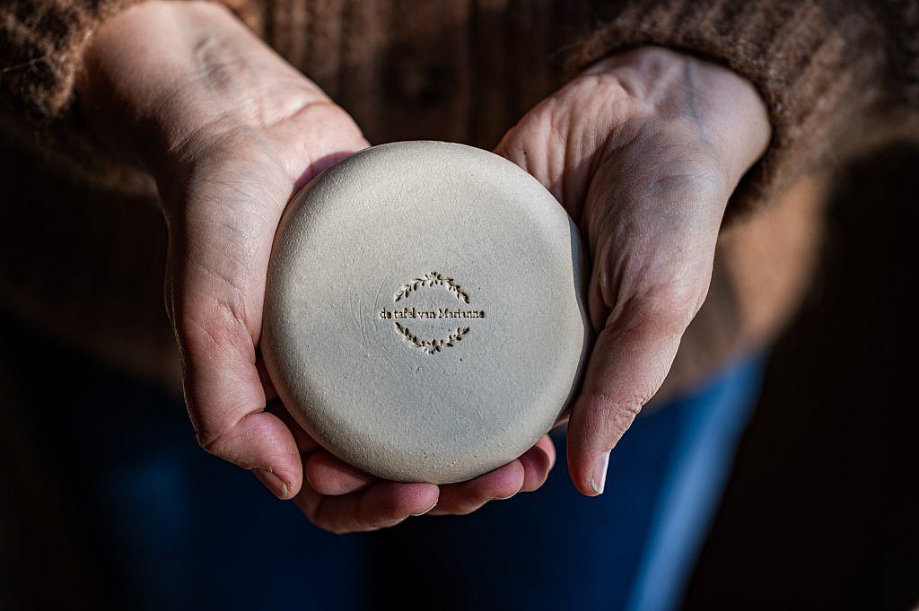 Vrouwenhanden houden een klein bordje in klei omgekeerd vast, je leest de indruk 'de tafel van Marianne'.