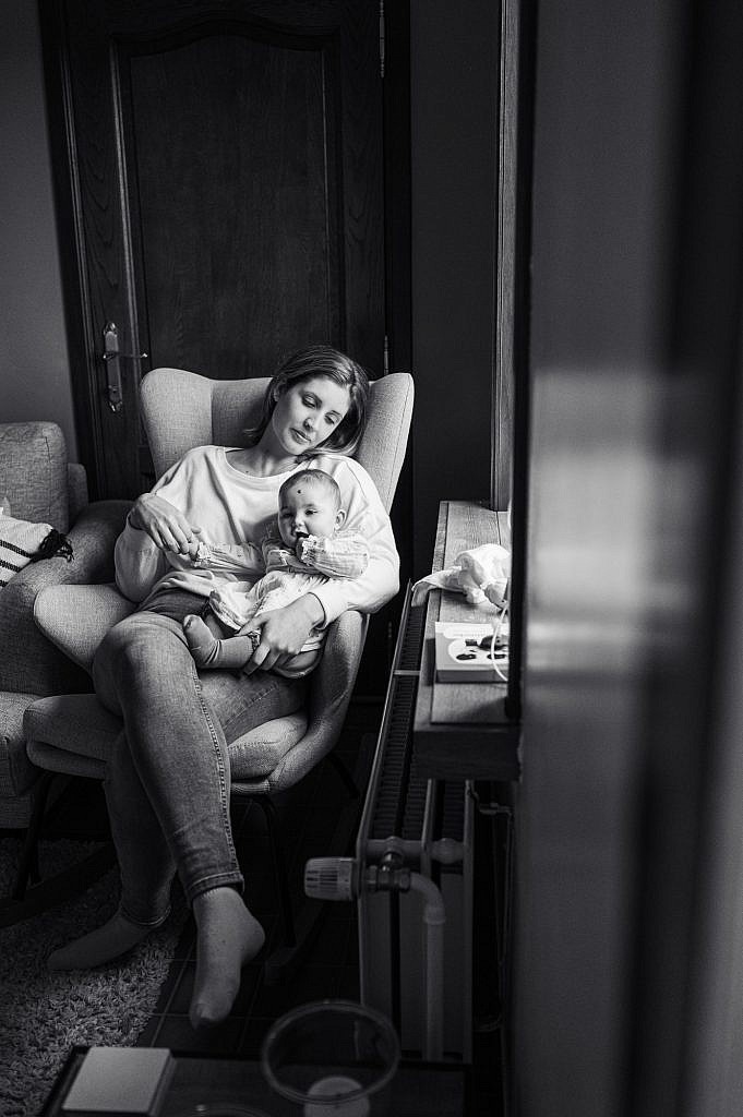 Zwart wit portret van mama met baby op de schoot in een zetel bij het raam.