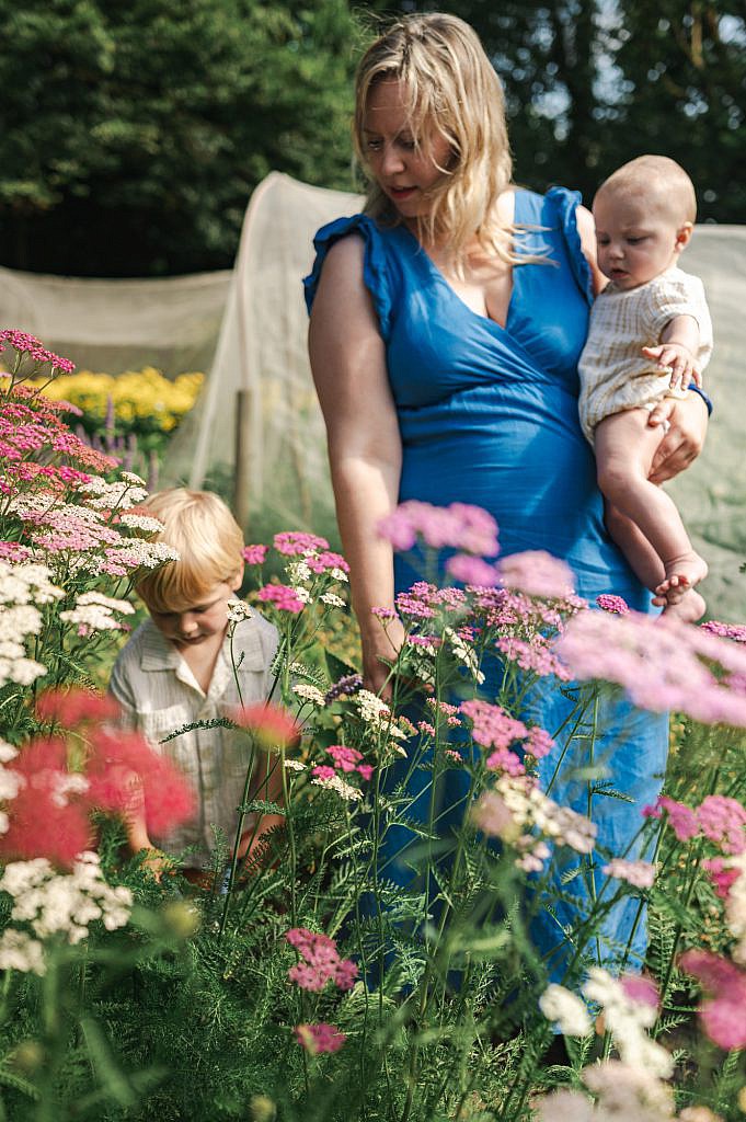 Mama in blauw kleedje met baby op de arm tussen een bloemenveld.