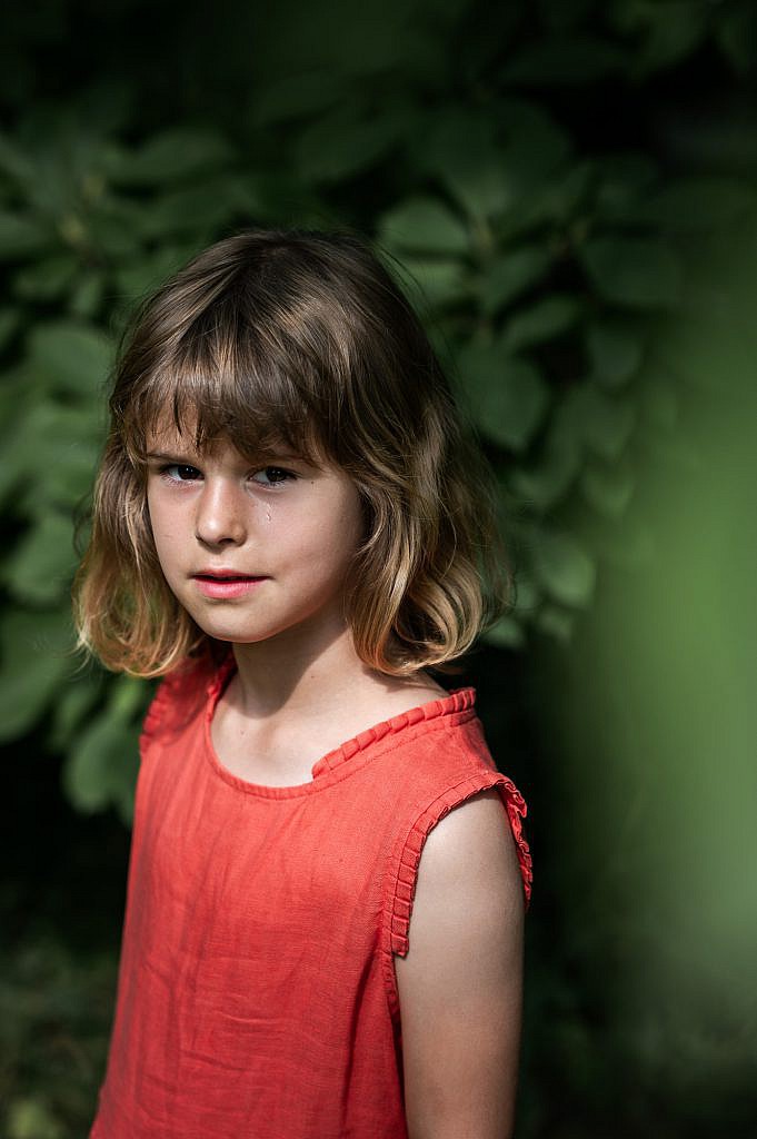Creatief portret van een klein, blond meisje met een rode jurk aan en een traan op haar wang die glimlachend in de camera kijkt.