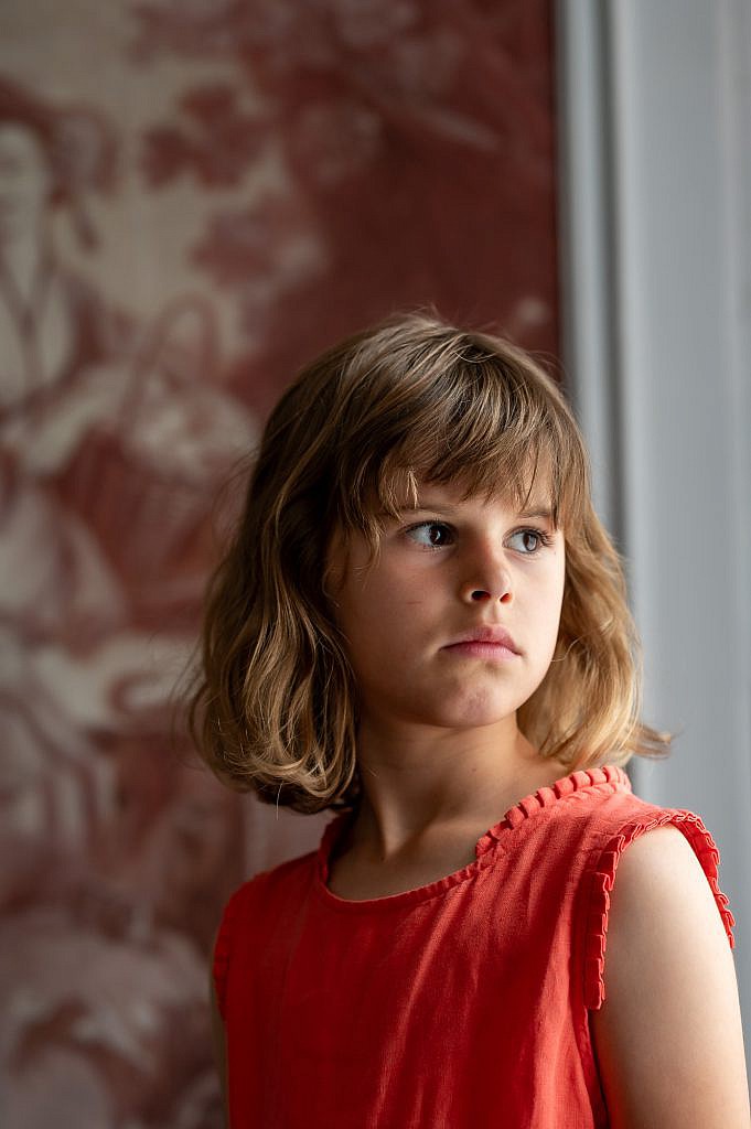 Creatief portret van jong meisje met blond krulhaar en rode jurk kijkt dromerig door het raam.