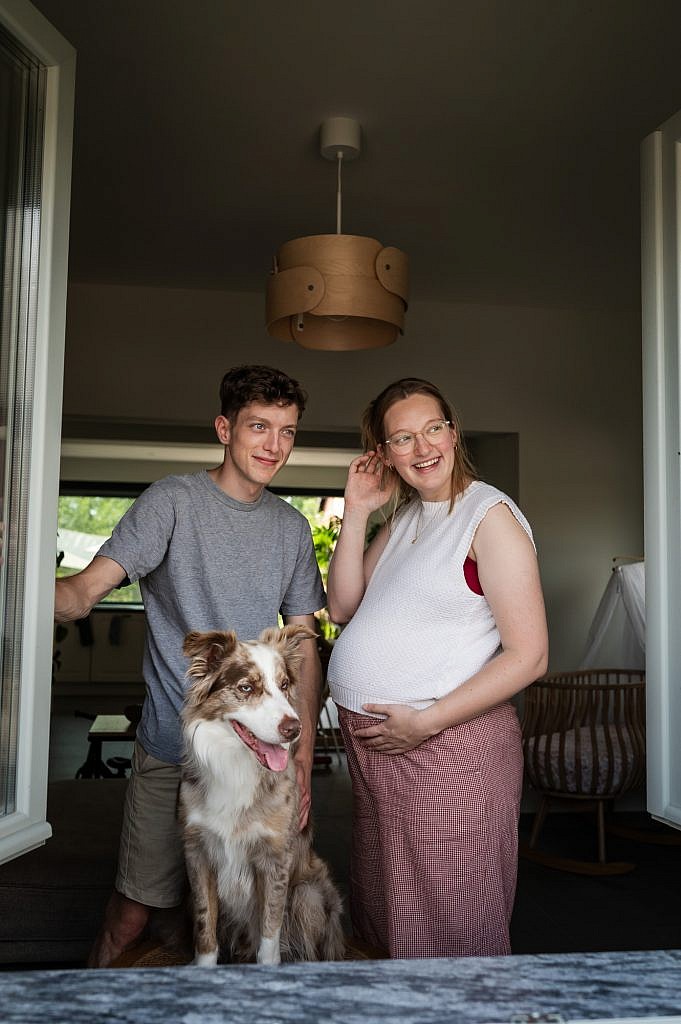 Zwangere mama met partner en hond kijken gelukkig door het raam, vastgelegd door fotograaf Nele Martens.